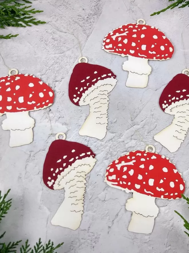 6 red and white paper mushroom decorations laying on a pale grey background.  Three of the toadstools have a bright red and white wide cap with a white stalk.  The other three toadstools have a dark cherry red and white pointy cap with a white stalkChristmas Mushroom Ornaments