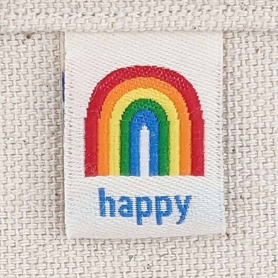 A close-up of a woven fabric tag attached to a textured off-white canvas material. The tag features an embroidered rainbow arch in red, orange, yellow, green, and blue, with the word 'happy' stitched in blue beneath it.