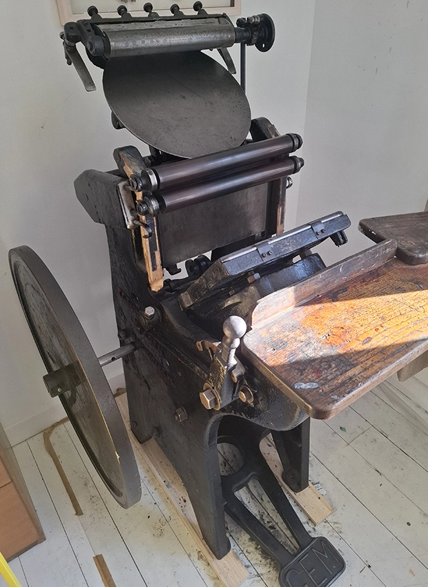 Anna Thundercliffe's printing press.  An antique printing press stands on white floorboards with sunshine slanting across it.