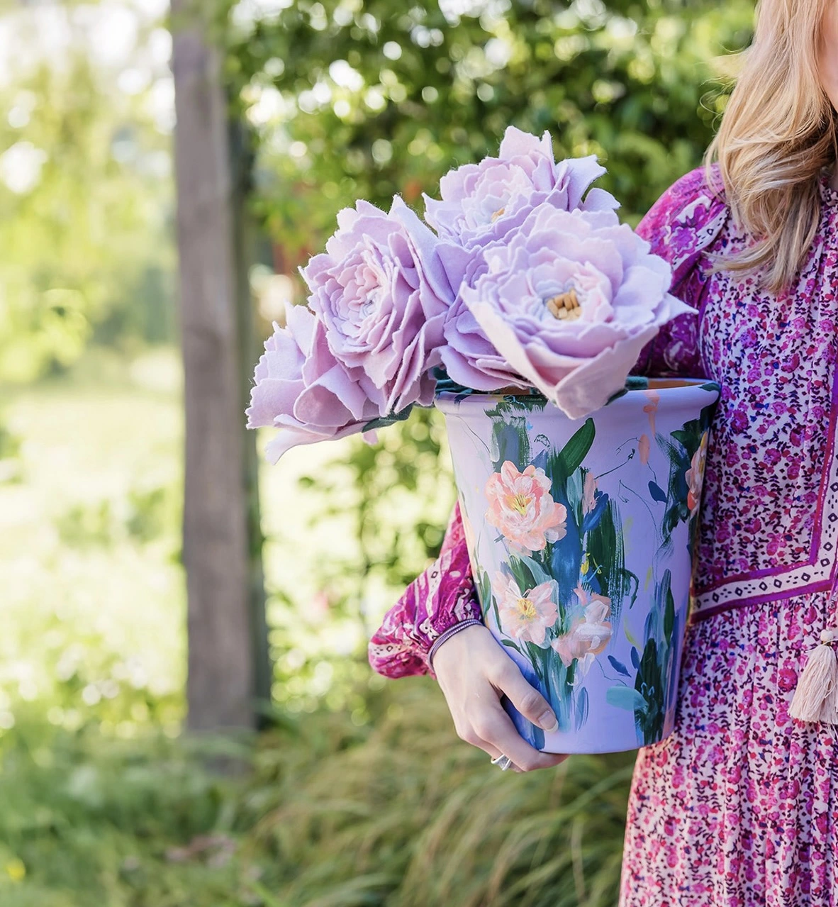 Hand painted floral lilac plant pot. A person in a pink floral dress holding a lilac plant pot