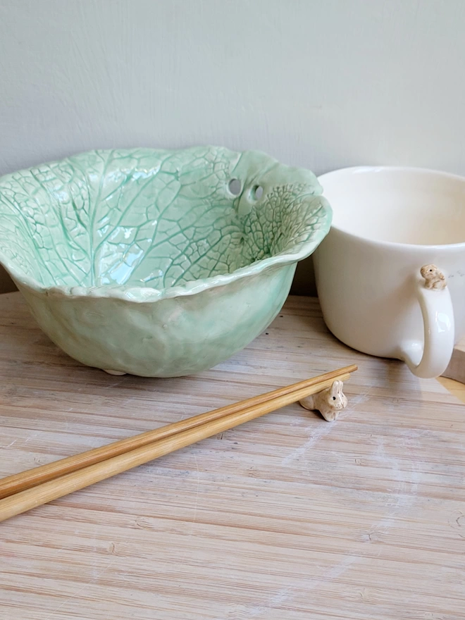 green ceramic bowl with holes for chop sticks and imprinted letuuce or cabbage leaves with chop sticks in front resting on a bunny holder and white bunny cup 