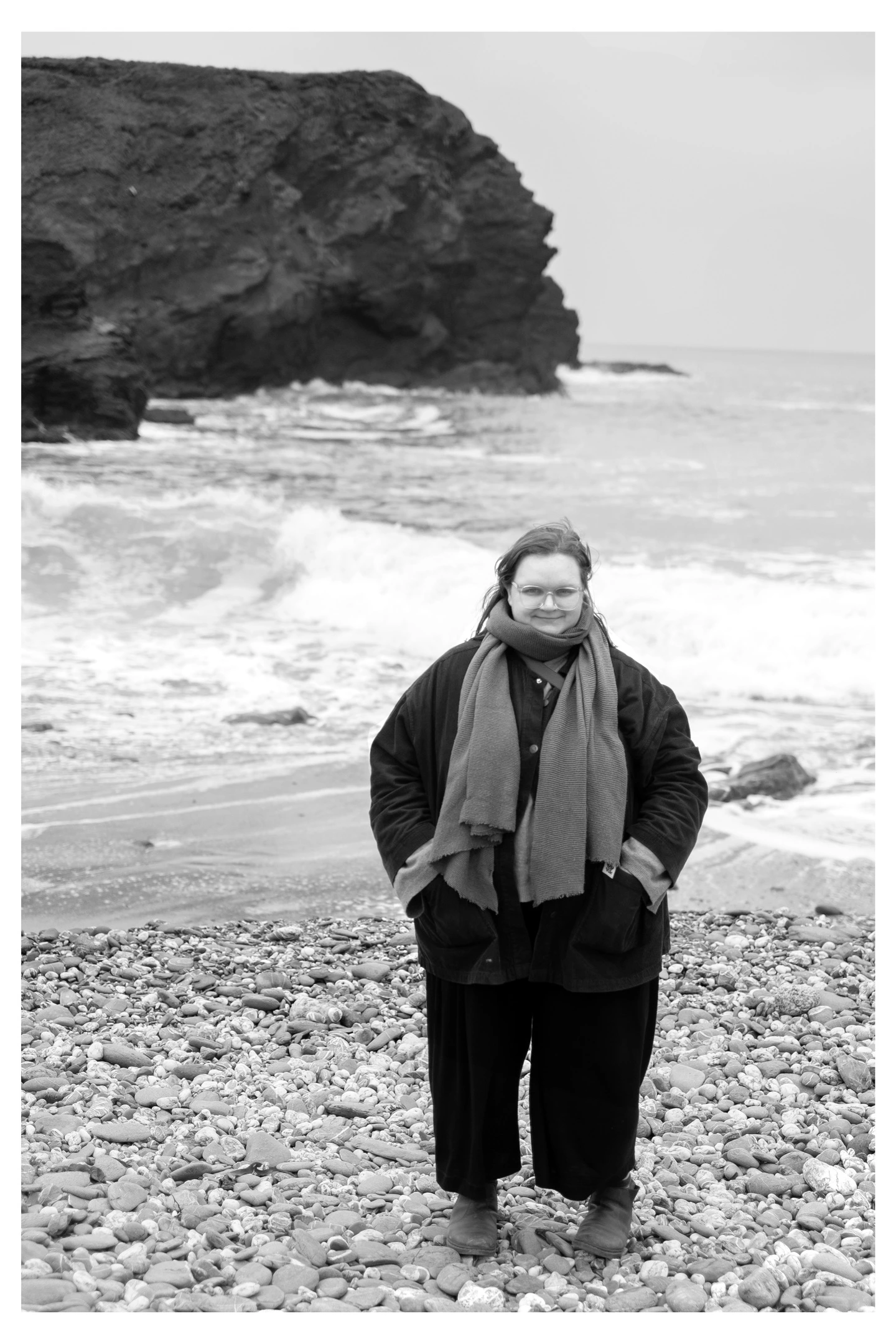 A black and white photograph of Meg, a white woman wearing a scarf, cord jacket and trousers stood on the beach
