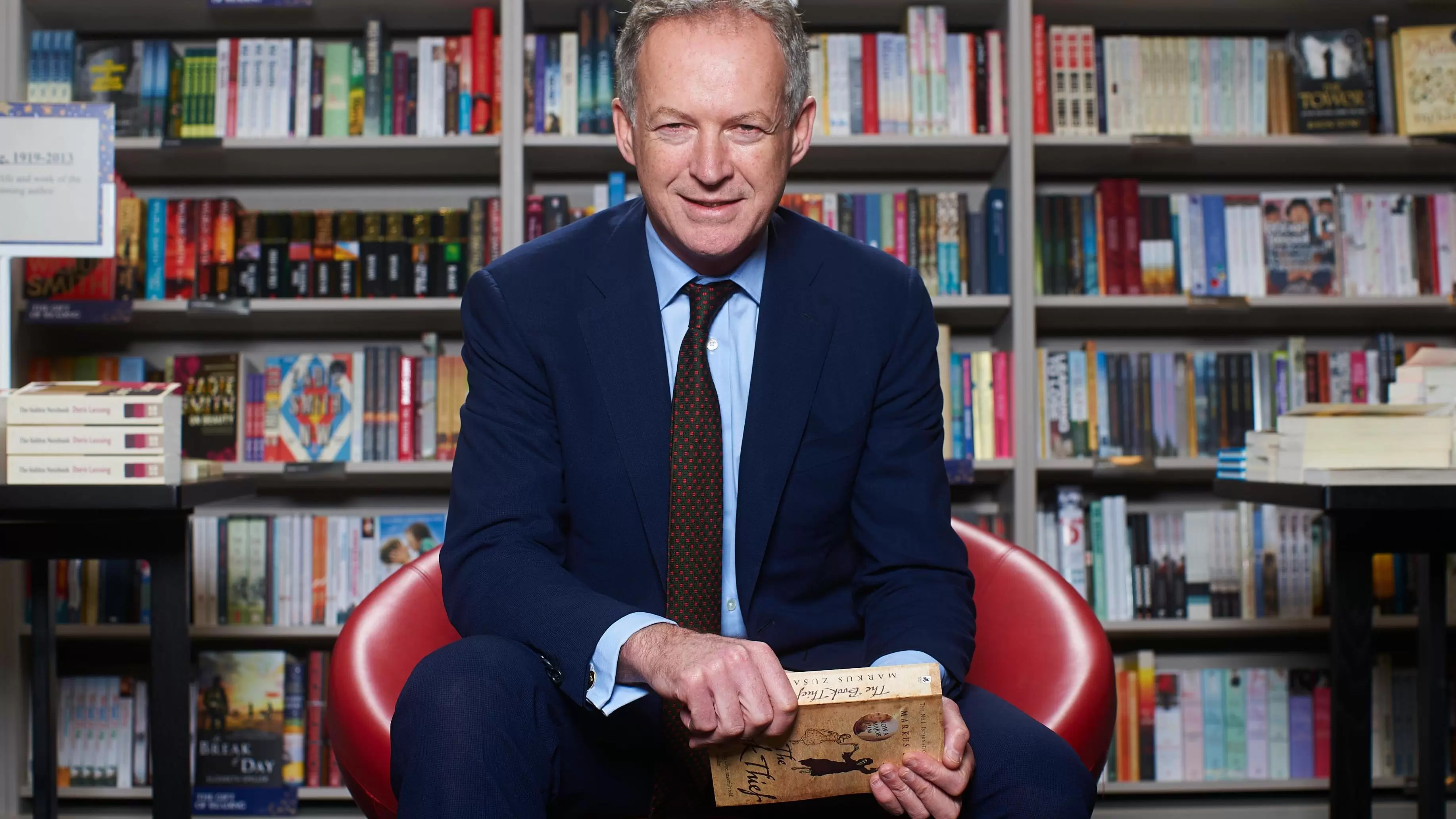 James Daunt, founder of Daunt Books, smiling at the camera holding a book, sat infront of a large bookshelf.