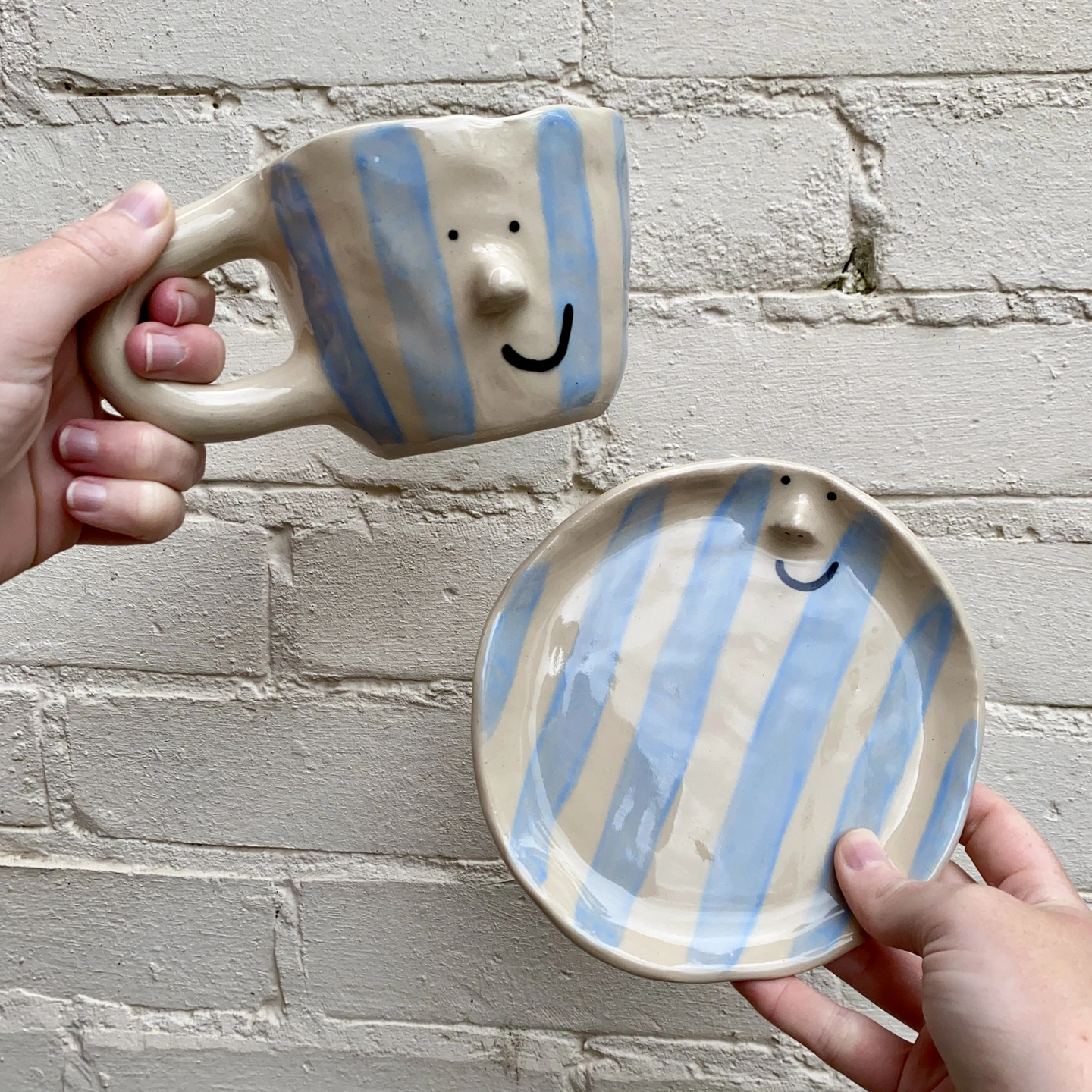 A photograph of a mug and a plate being held infront of a cream brick wall. Both the mug and plate have pale blue stripes, on a natural clay coloured background. With black eyes and smile painted, with a 3D nose.