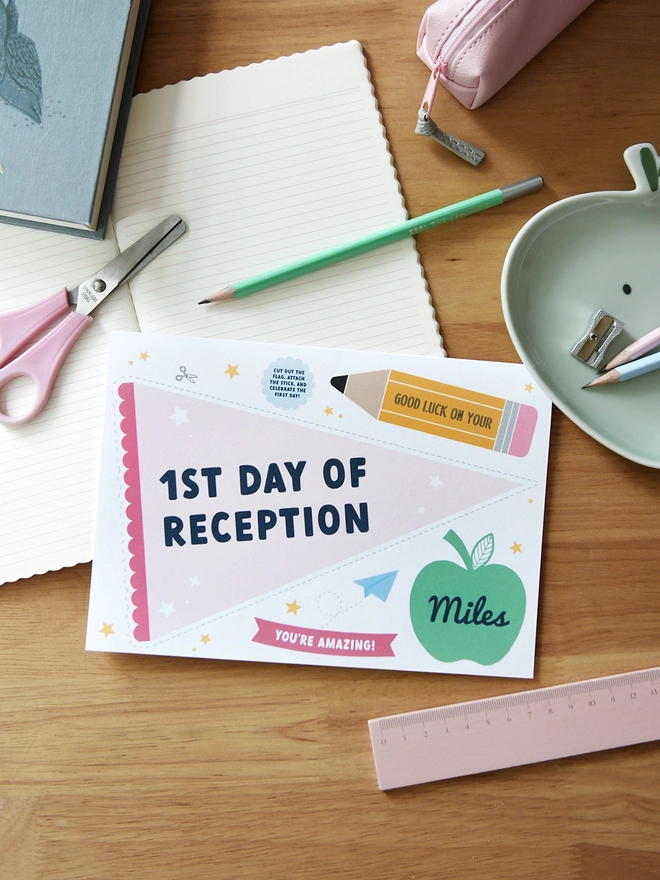  A "1st Day of Reception" card lies on a wooden desk surrounded by school supplies like scissors, pencils, a ruler, and a pencil case. The card includes a personalised name, encouraging messages, and playful designs.