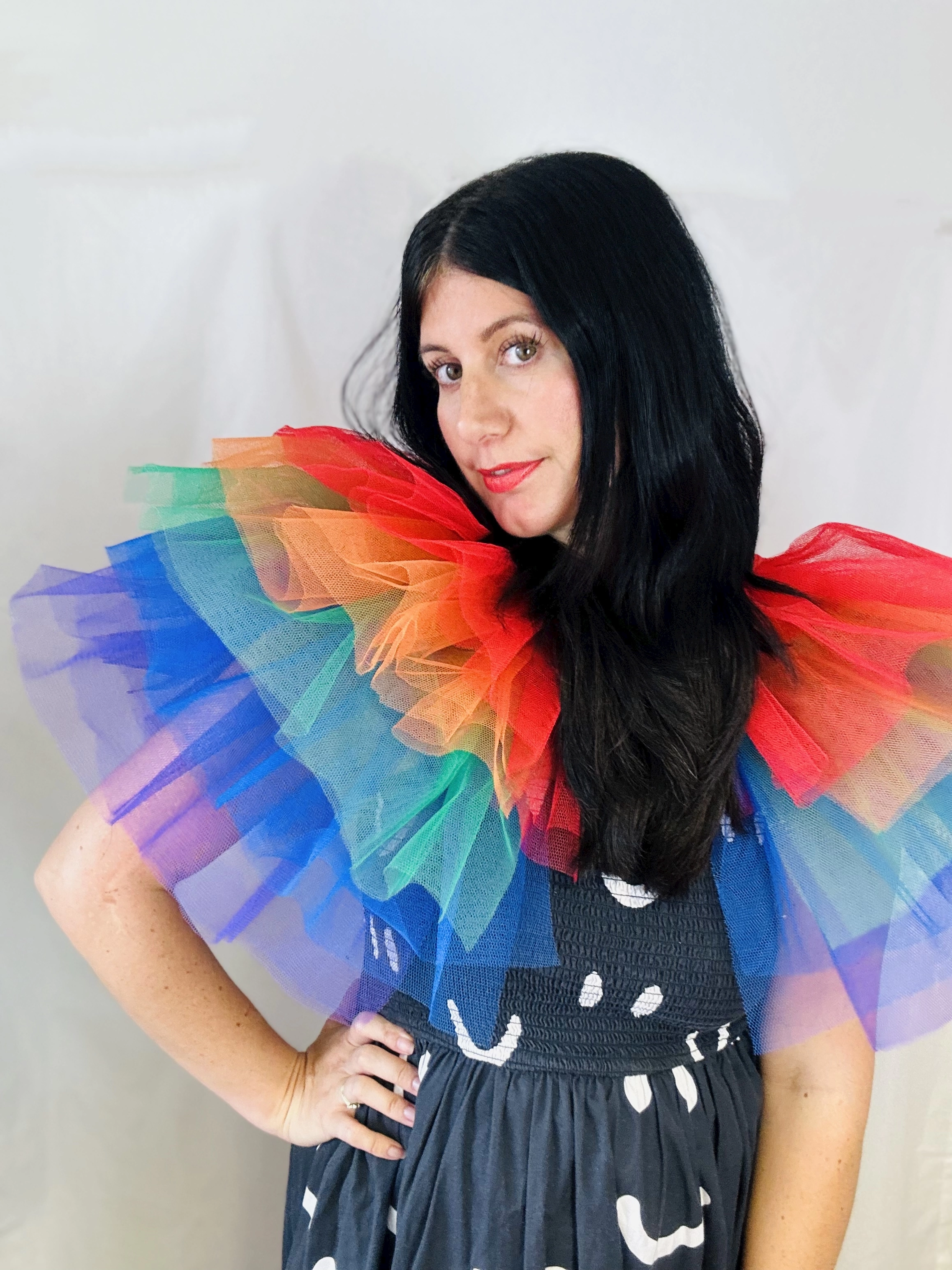 Susie, Female Founder Designer and Maker at Let Us Pretend wears a rainbow cape, she has black hair and pale skin and wears red lipstick and a black and white dress