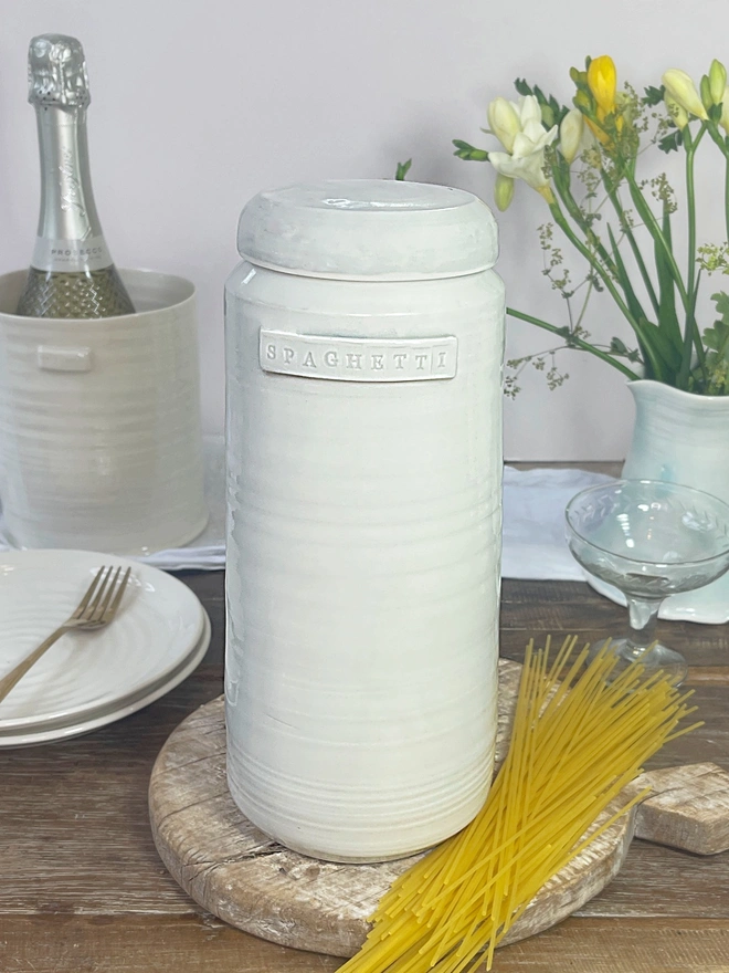 Spaghetti storage jar in grey glaze sat on a table with dried spaghetti, a jug or flowers, some wine in a wine holder and plate