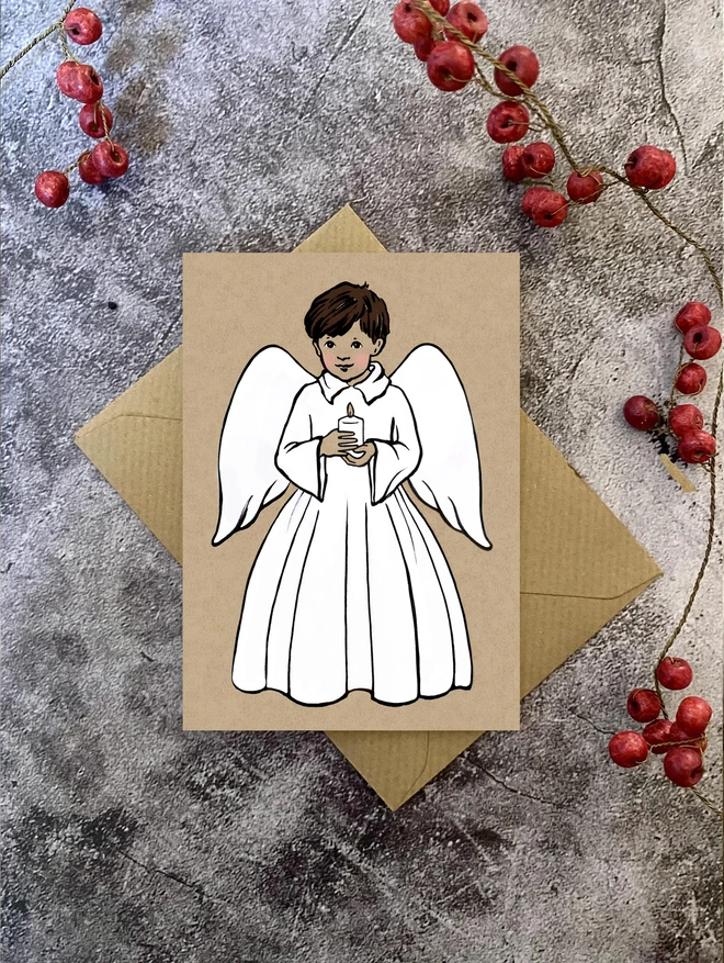 greeting card of a dark haired boy angel holding a candle laid on a Kraft envelope with red Christmas berries