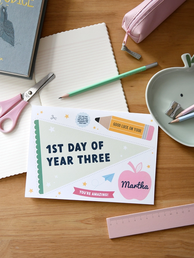  A "1st Day of Year Three" card lies on a wooden desk surrounded by school supplies like scissors, pencils, a ruler, and a pencil case. The card includes a personalised name, encouraging messages, and playful designs.