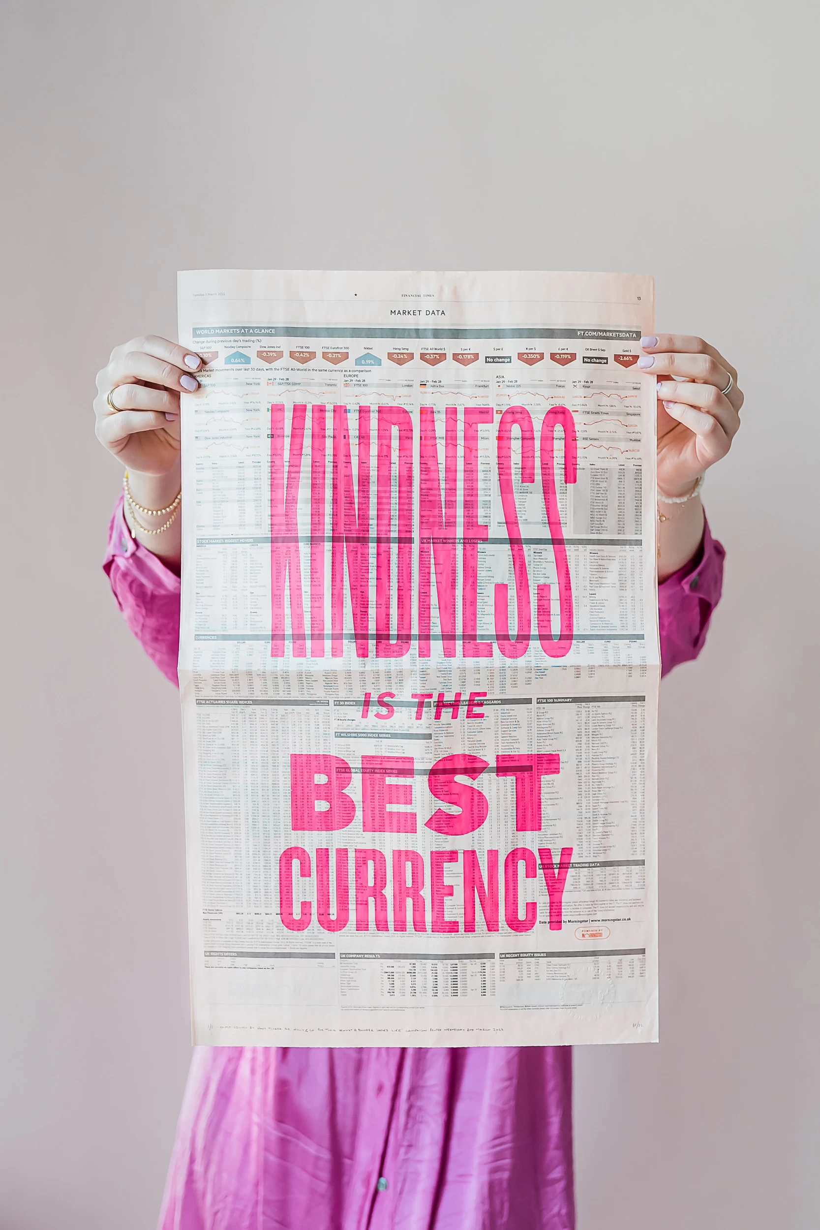 Woman holding up a sheet of newspaper with the words Kindness is the best currency written on it in pink