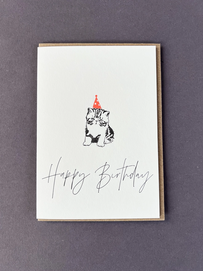 A cute little kitten in a neon orange party hat with "Happy Birthday" beautifully written in a modern calligraphy underneath.