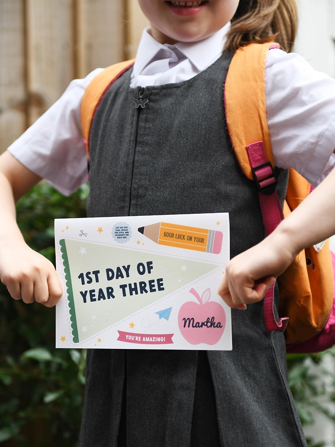 A young child, wearing a school uniform and backpack, holds a "1st Day of Year Three" card with excitement. The card features encouraging messages and playful designs, perfect for celebrating a child's first day at school.