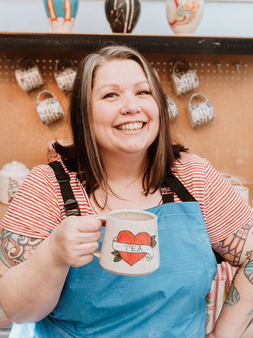 Emma Puddick, a white woman in her forties looks directly at camera smiling.  She is wearing a blue apron, and red and white striped shirt.  she is holding a handmade ceramic mug, with a heart and banner tattoo reading TEA