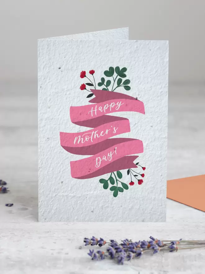 A white card stood up on a white wooden floor with lavender in front of it. The card has a floral design with 'Happy Mothers Day' on.