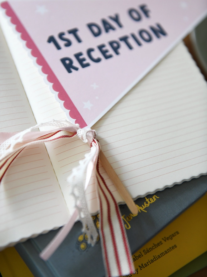 A pink pennant flag with the words "1st Day Of Reception" lays on an open notebook on a wooden desk. Various stationery items surround it.