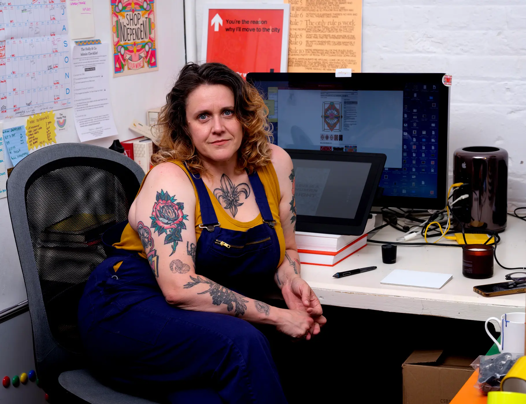 Picture show Rebecca sitting at her studio desk in Margate. There is a computer and drawing tablet just behind her and colourful pictures stuck to the white walls. On her desk is a pen, a candle, a phone and mousebad. Rebecca is wearing a yellow vest and royal blue dungerees. She has curly brown and blonde hair and many colourful tattoos.