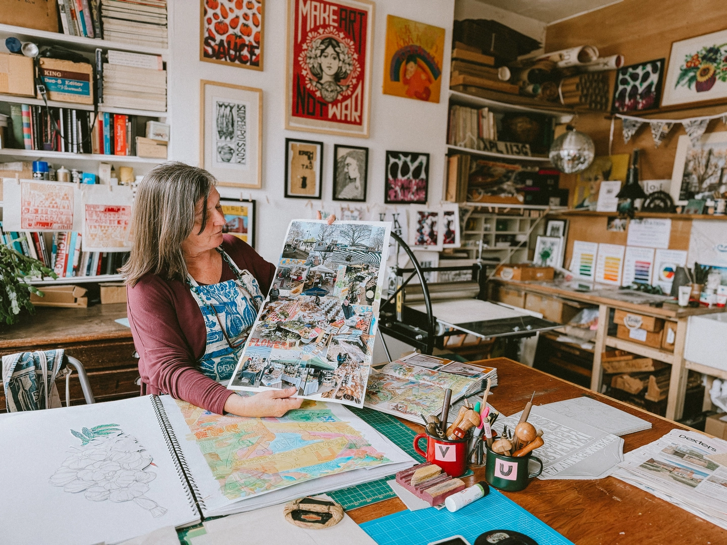 Printmaker Kate Guy in her London art studio holding up a detailed illustrated print of Kew Village Market over open sketchbooks on her workbench.