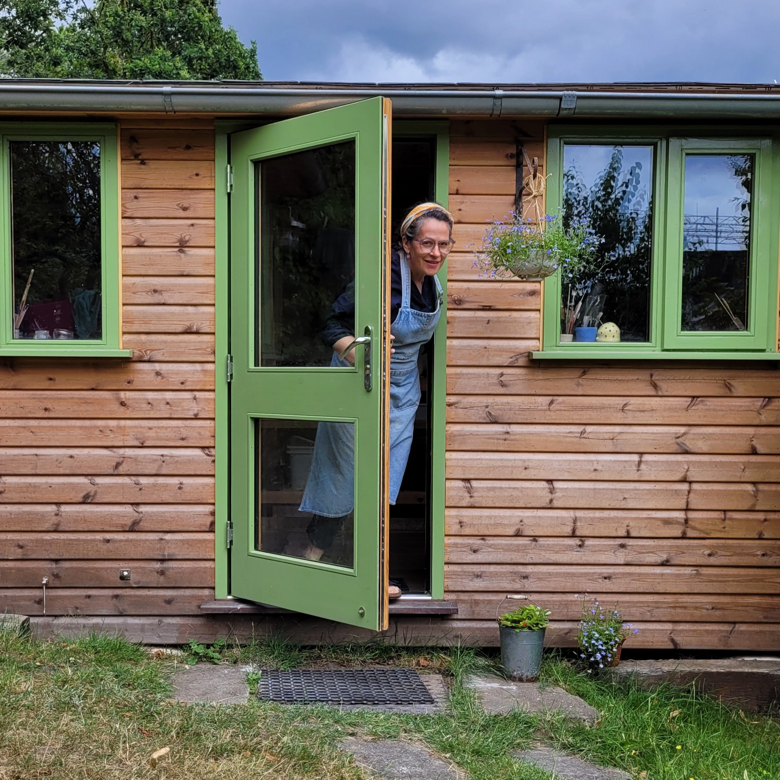 lucyandjaneceramics Lucy is standing in the doorway entrance to her garden pottery studio a wooden cabin with green windows and door 
