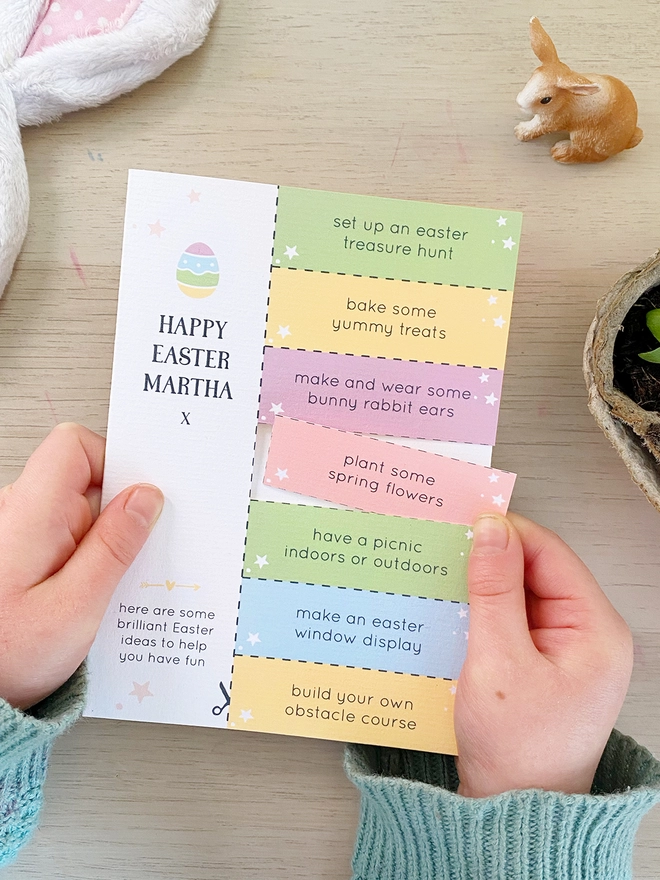 A personalised Easter greetings card with seven cut out activity coupons is being held by a young child above a wooden desk.