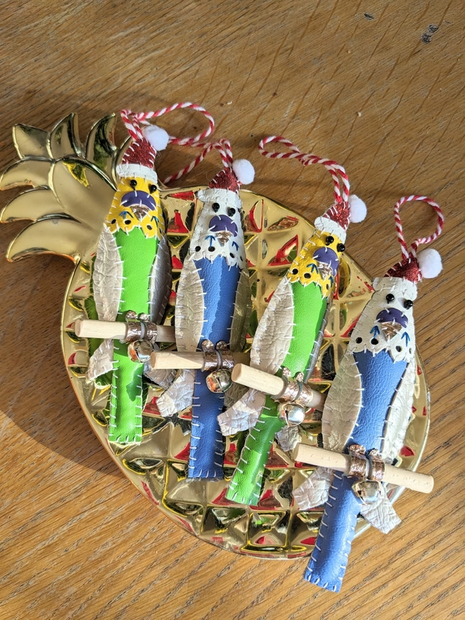 A gold pineapple dish containing four hand stitched faux leather budgie Christmas decorations wearing Santa hats. Two birds are blue, two are green.