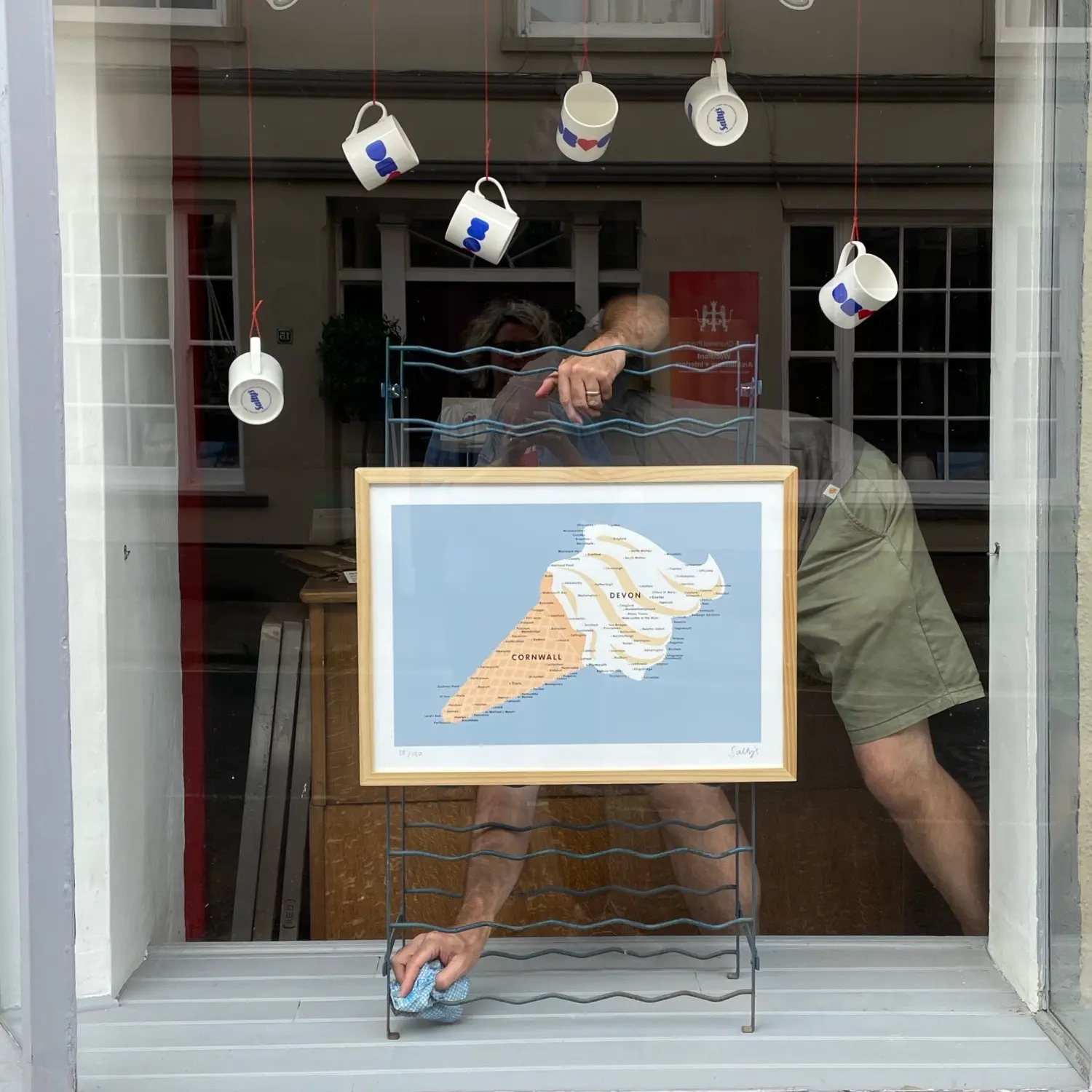 A man dusts a shop window, obscured by a wire frame holding up a picture of Devon and Cornwall in the shape of an icecream. China mugs are suspended above him, each with a design on saying Devon. 