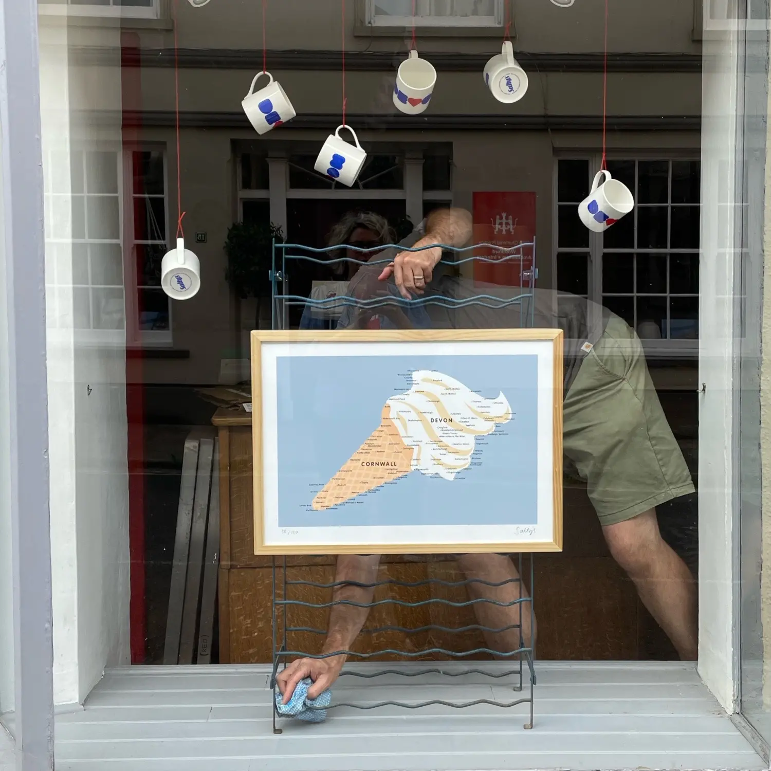 A man dusts a shop window, obscured by a wire frame holding up a picture of Devon and Cornwall in the shape of an icecream. China mugs are suspended above him, each with a design on saying Devon.
