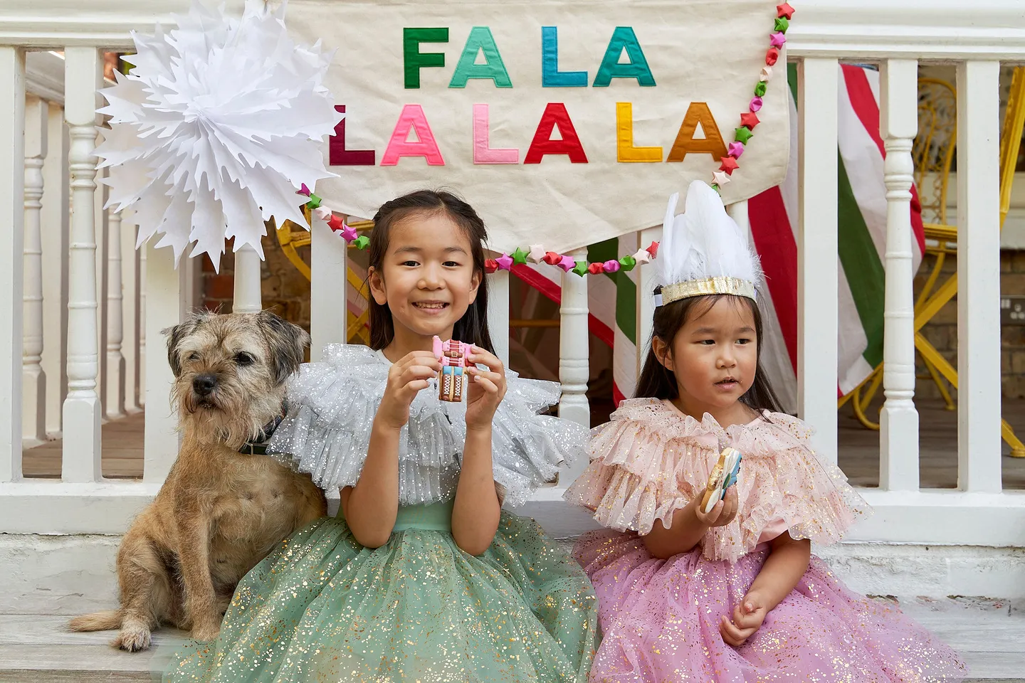 Kids dressed up in sparkly Christmas dresses eating a nutcracker biscuit with a fa la la la colourful banner behind them