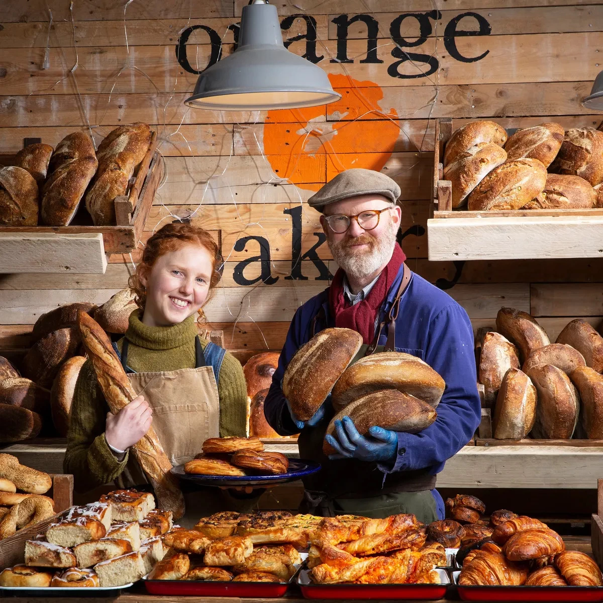 Kitty Tait and Al Tait, founders of Kitty's Kits and The Orange Bakery, smiling at the camera, stood in their bakery, holding bread.