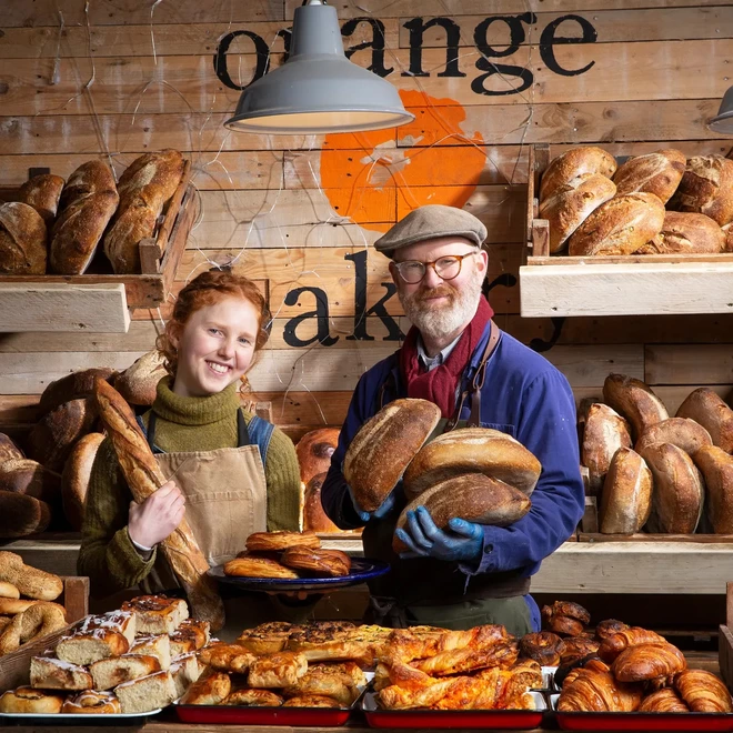 Kitty Tait and Al Tait, founders of Kitty's Kits and The Orange Bakery, smiling at the camera, stood in their bakery, holding bread.