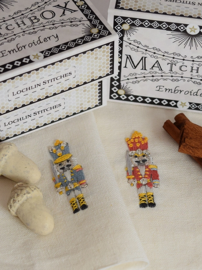 A mini embroidered nutcracker,  embroider in red or blue - colours are interchangeable. Displayed with decorative acorns and flora.