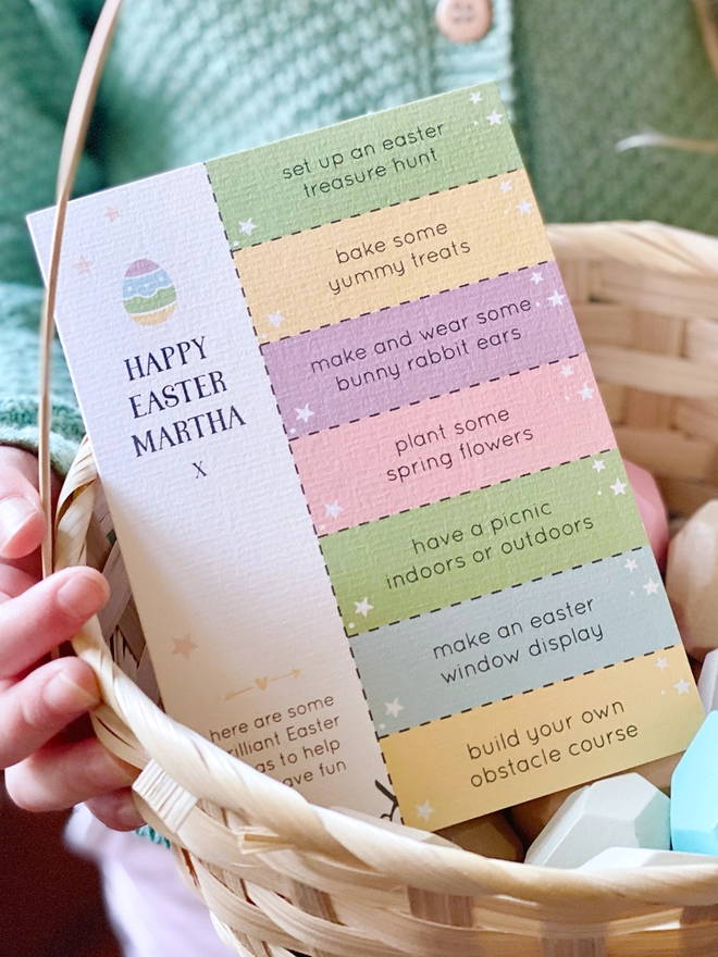 A personalised Easter greetings card with seven cut out activity coupons is tucked into an Easter basket being held by a young child.