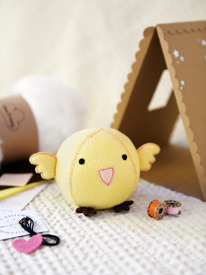A handmade yellow felt chick toy sits beside the craft kit contents used to make it and a cardboard house.