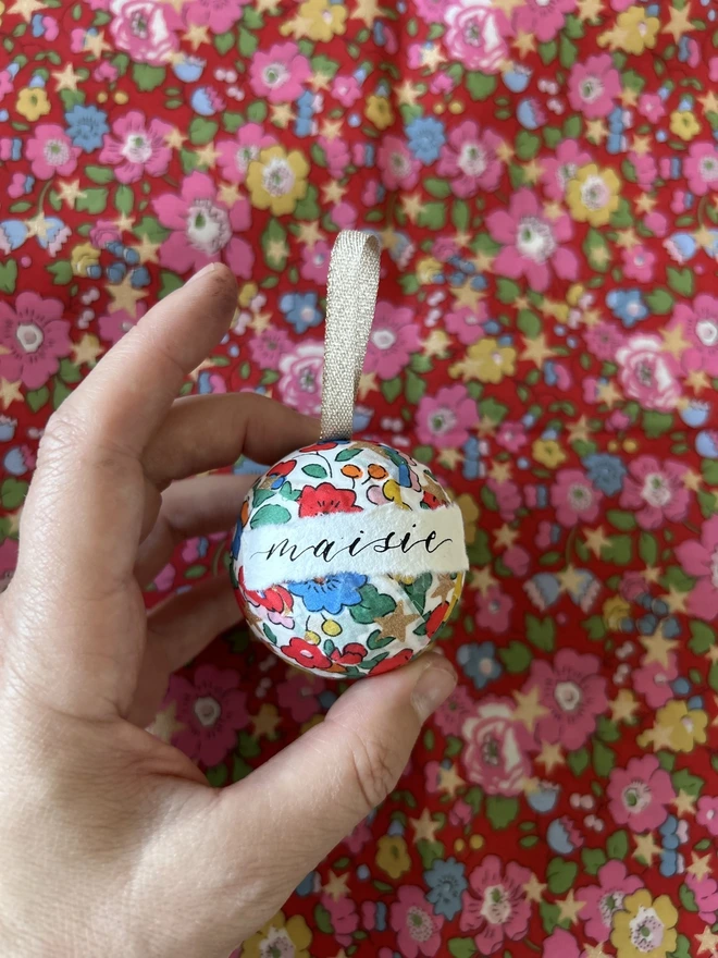 Mini Liberty fabric Christmas bauble with personalised Maisie label held in front of a Liberty fabric backdrop 