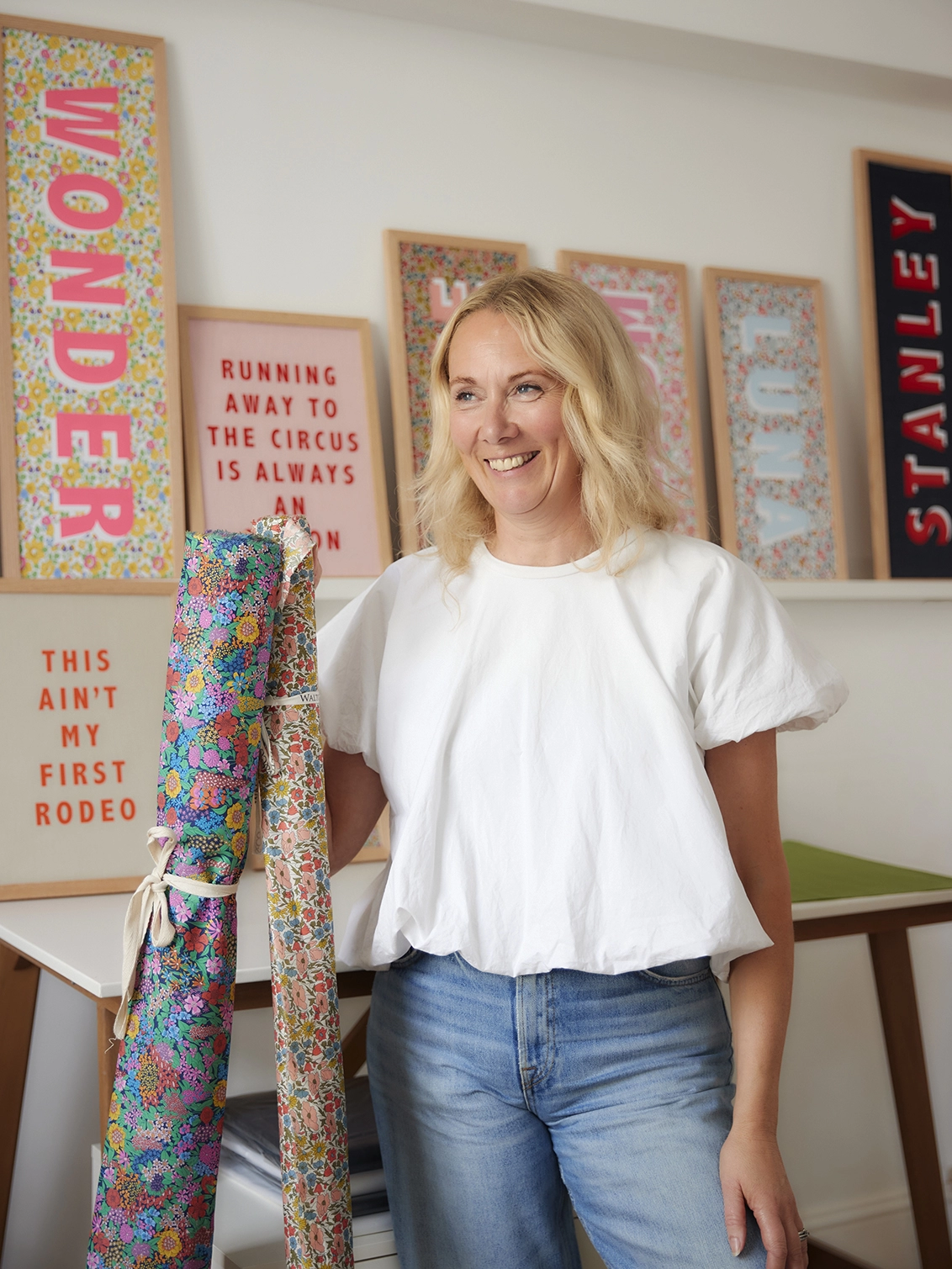 Catherine Colebrook in her studio holding 2 rolls of Liberty fabric, with Liberty word/name pictures in the background.