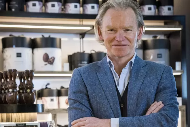 Angus Thirlwell, co-founder of Hotel Chocolat, smiling at the camera with his arms crossed, stood infront of a wall of Hotel Chocolat products.  