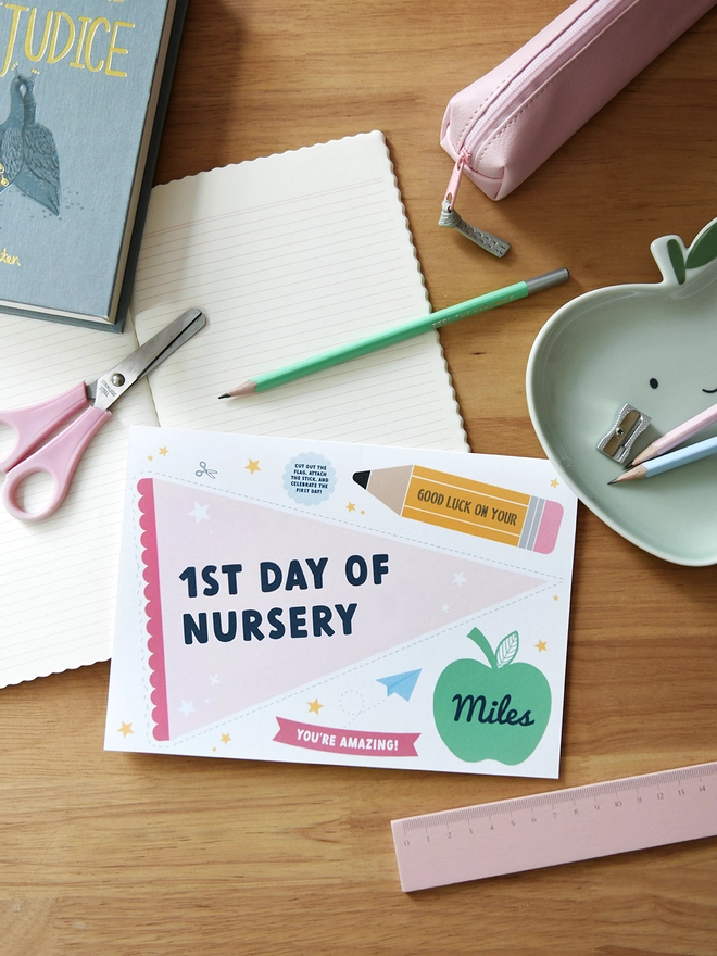  A "1st Day of Nursery" card lies on a wooden desk surrounded by school supplies like scissors, pencils, a ruler, and a pencil case. The card includes a personalised name, encouraging messages, and playful designs.