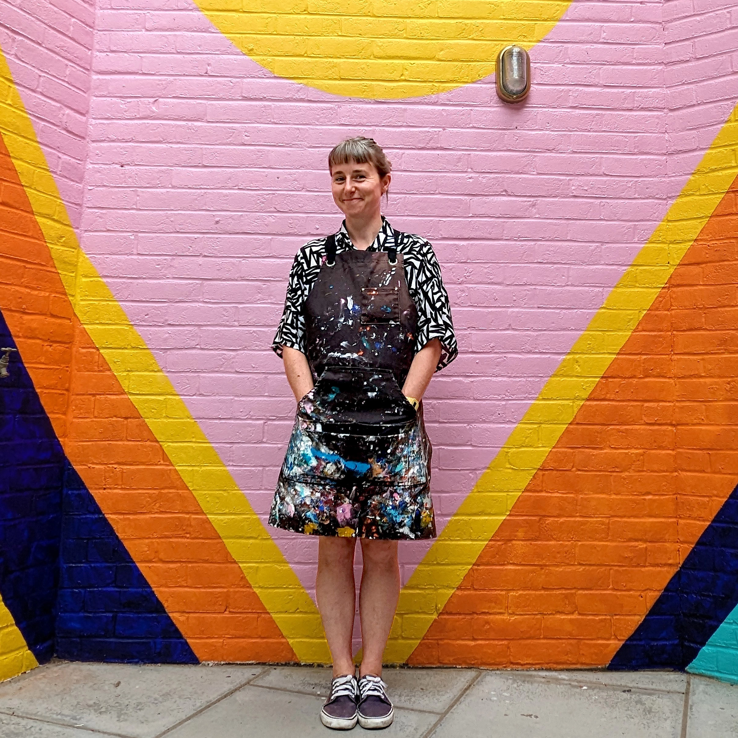 Artist Survival Techniques stands in front of a mural wall painted with a large pink triangle along with yellow, orange and blue and a yellow sun above