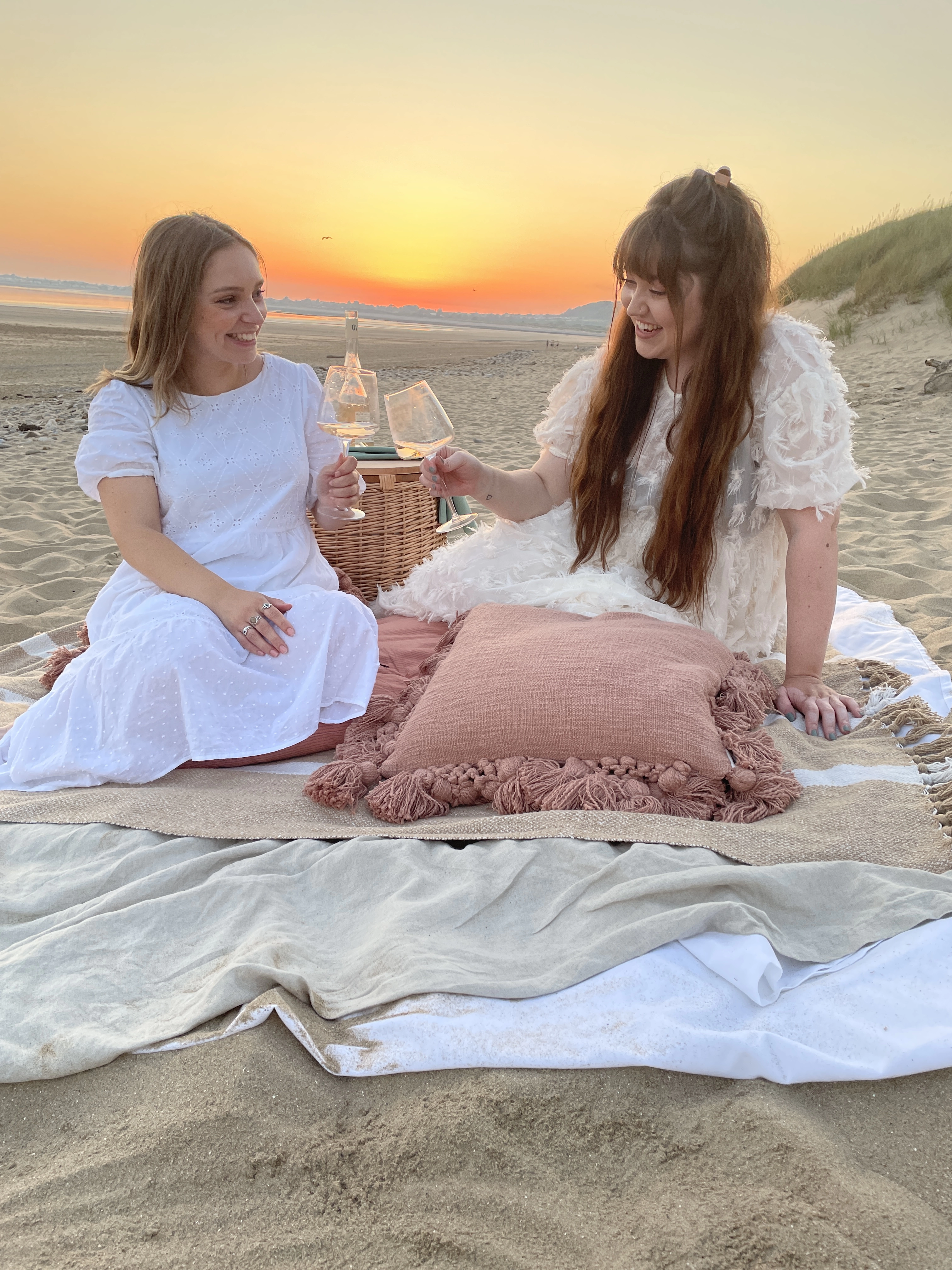 the two female founders of alex and annie studio sit on a beach with blankets and a picnic cheers'ing with glasses of wine