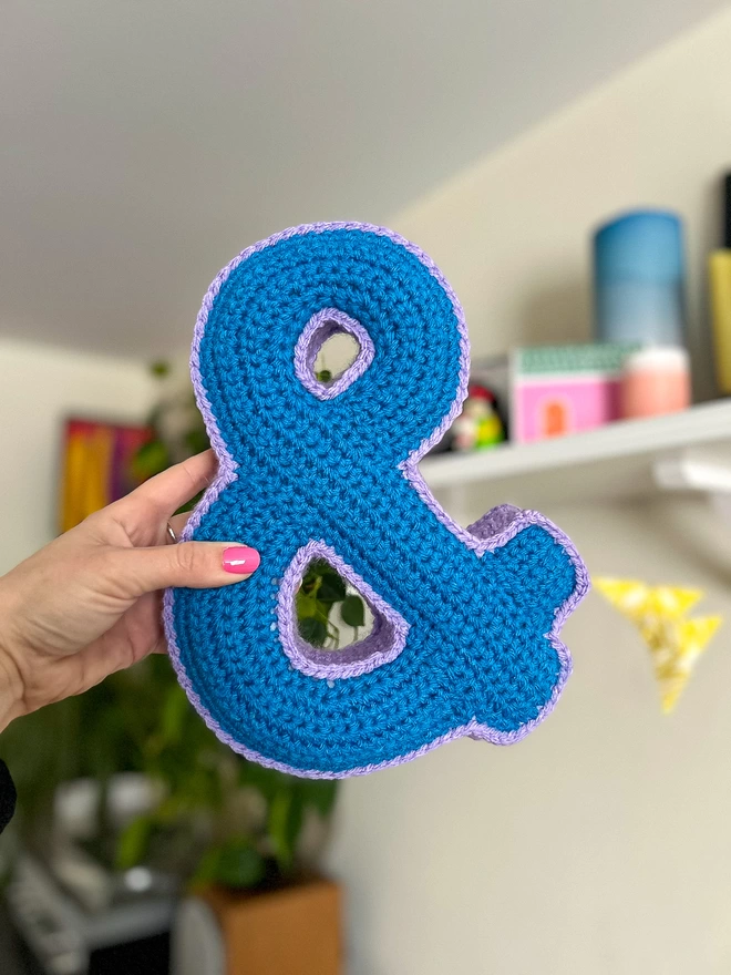 A hand holds a Crochet Cushion shaped like an Ampersand in Blue and Lilac