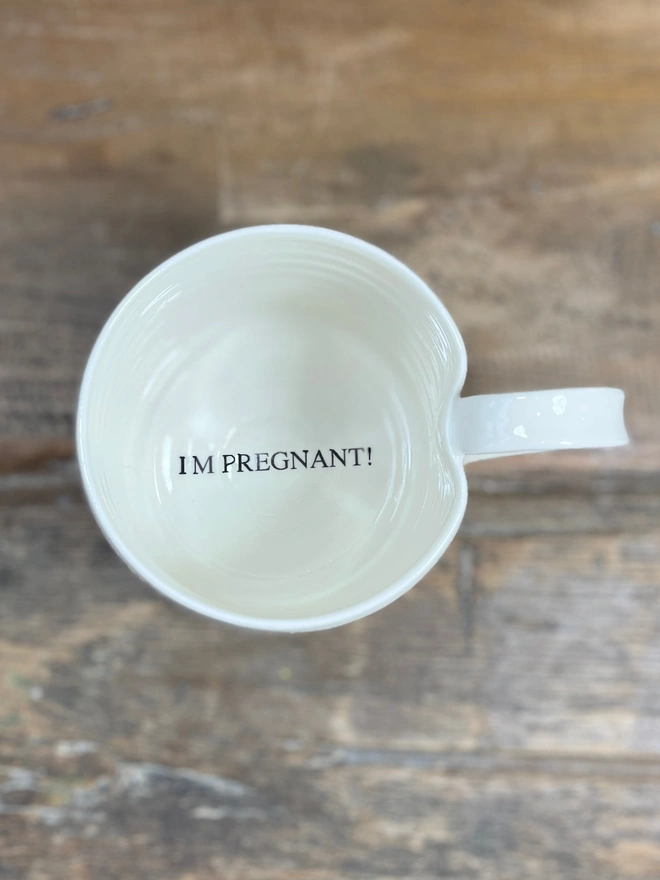 Mug with 'I'm Pregnant!' announced in black text on the inside base. Mug is satin cream glazed and is sat upon a rustic wooden table top. 