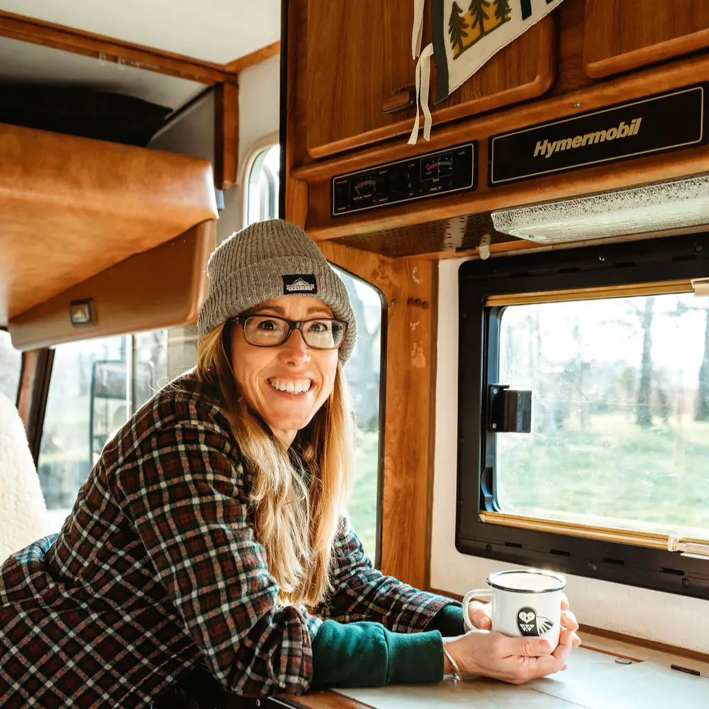 A lady wearing a check shirt and beanie hat leaning on the sideboard of a campervan with an emamel mug