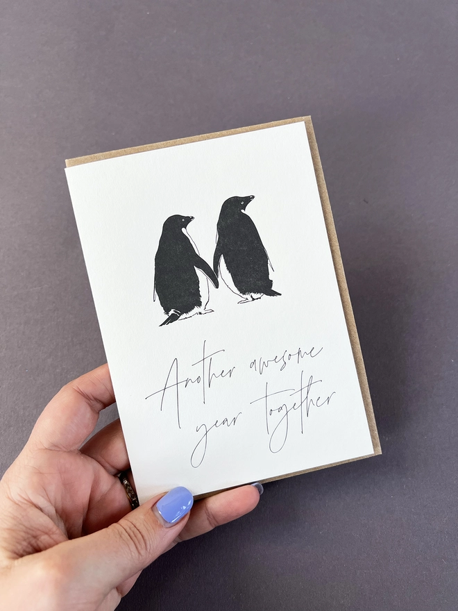 The back of two penguins lovingly holding flippers with "Another awesome year together" written beautifully in a modern calligraphy.