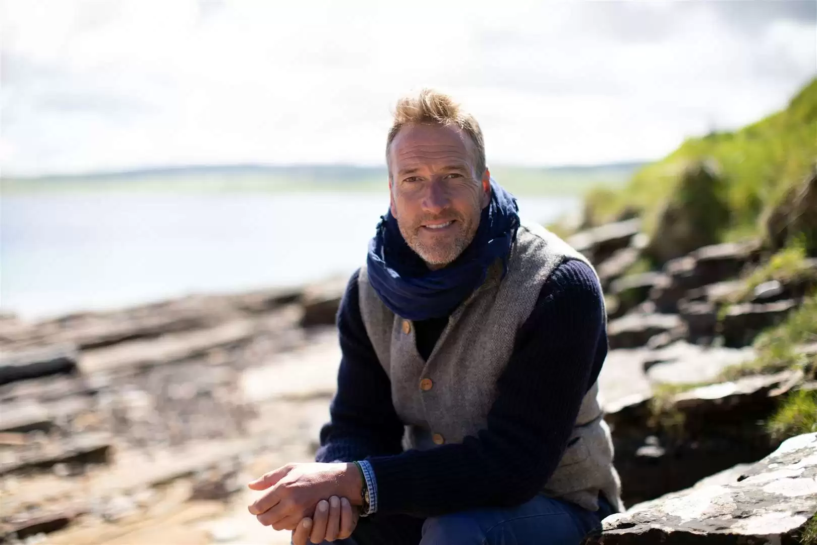 Ben Fogle, broadcaster, writer & adventurer, smiling at the camera sat on a beach.