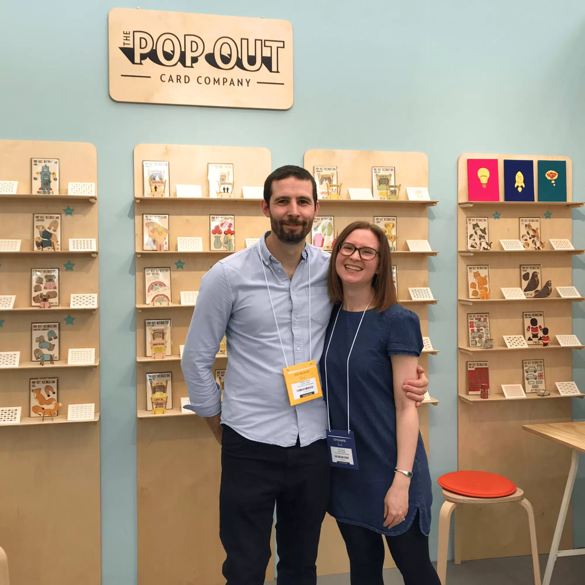 Jonathan and Louisa Aspinall of The Pop Out Card Company hugging and standing in front of their 3D laser cut pop out cards on display at a trade show