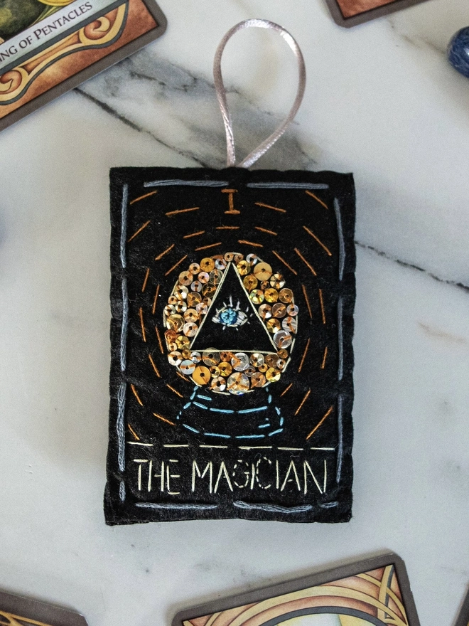 A handcrafted felt ornament with sequins and embroidery depicting "The Magician" tarot card. Colourful embroidery with an eye inside a crystal ball. 