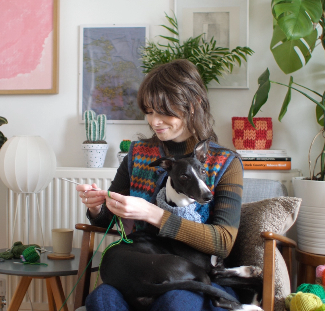 Emily sitting in her colourful studio crocheting a cactus. Her whippet is sitting on her lap.