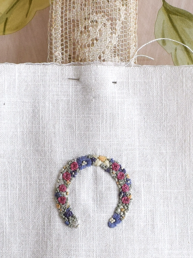 An embroidered Floral Meadow Horseshoe, of Lavender Blues and Buttermilk yellow blossoms.   Pinned to a wide lace ribbon.