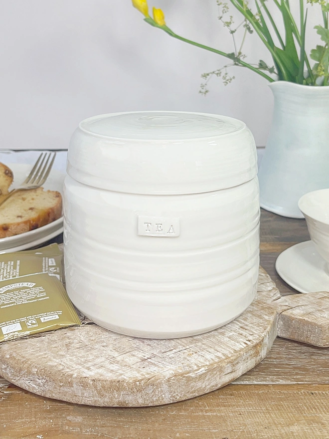 tea storage jar in cream porcelain with lid. Sits upon a wooden chopping board with tea bags and a slice of cake on a plate in the background. 