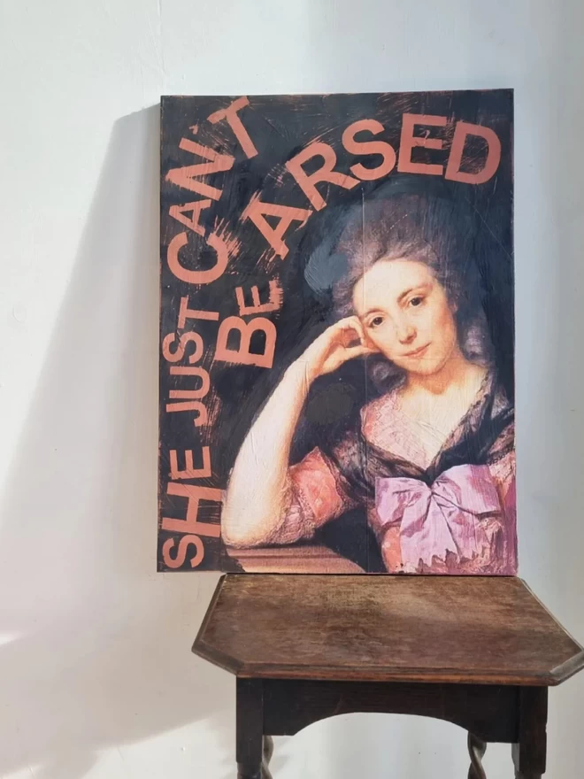 Painted ply wood art panel for the wall featuring an antique regal portrait of a lady. The colours are Peachy pink and black and the text reads in pink ‘she just can’t be arsed’.