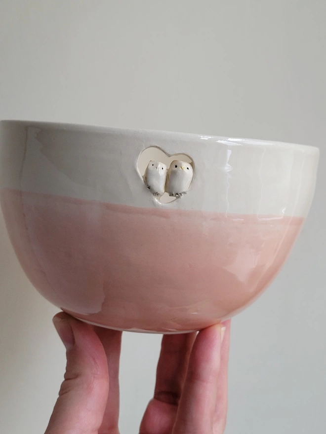 a pink and off white pottery bowl being held aloft in a hand with a couple of tiny ceramic owls sitting in a heart cut out 